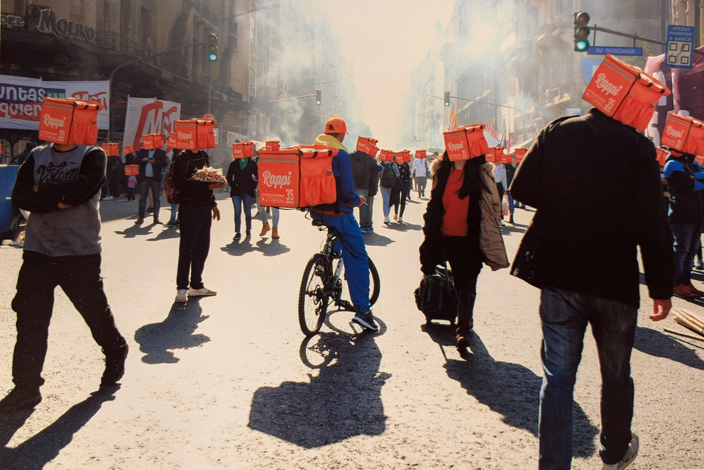 Viele Menschen befinden sich auf einer Straße, ihre Gesichter sind verdeckt von Essensauslieferungsboxen. 